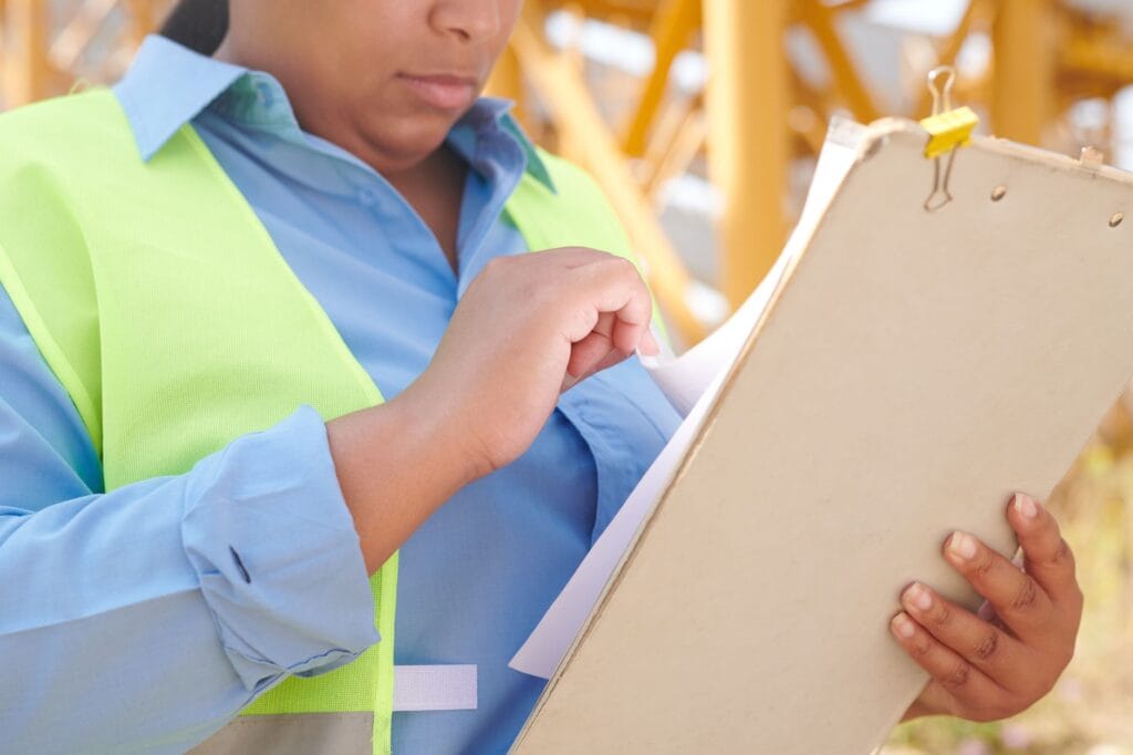A female engineer in safety gear reviewing documents on a clipboard at a construction site.