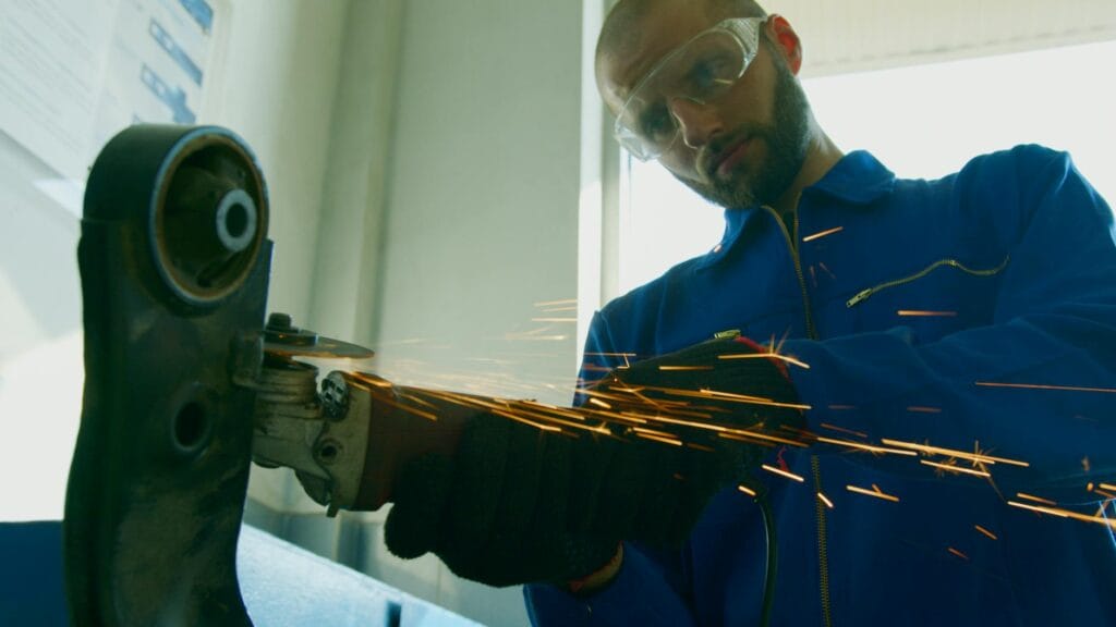 A mechanic wearing protective gear operates a grinder, creating sparks in a workshop setting.