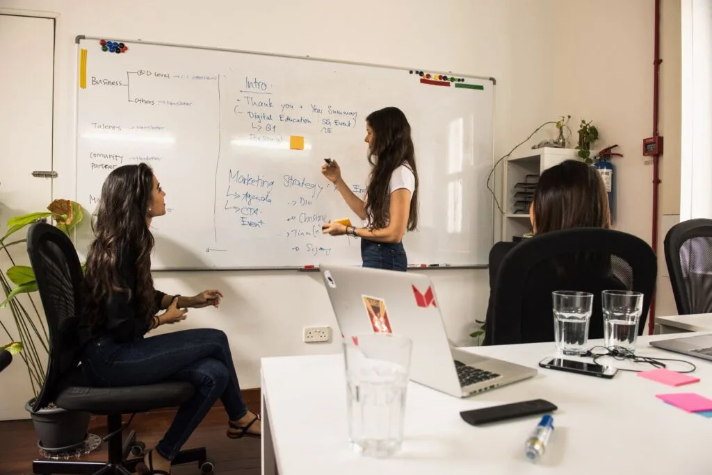 Business women discussing strategy at a whiteboard in a modern office setting.