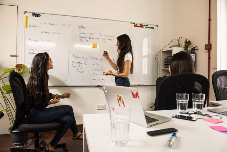 Business women discussing strategy at a whiteboard in a modern office setting.
