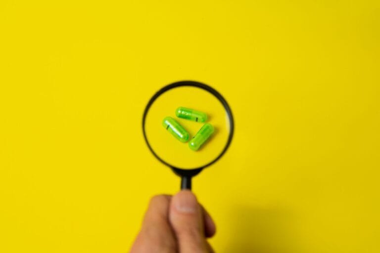 Top view of crop unrecognizable person holding magnifier over green pills placed on bright yellow surface in studio