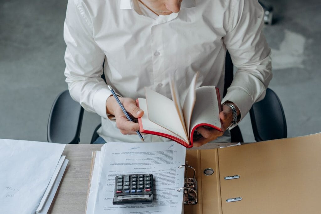 Man in white shirt reading book at office desk with calculator and documents.
